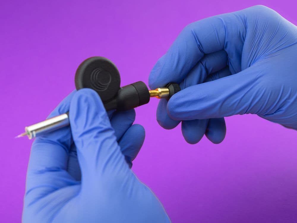 Gloved hands carefully assemble a small, metallic and black precision instrument on a purple background.
