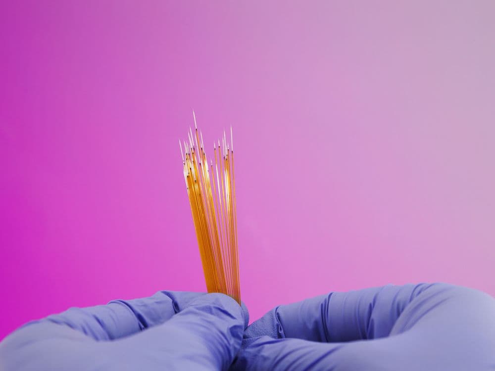 Gloved hands hold and stretch a viscous, amber-colored sticky substance against a pink background.